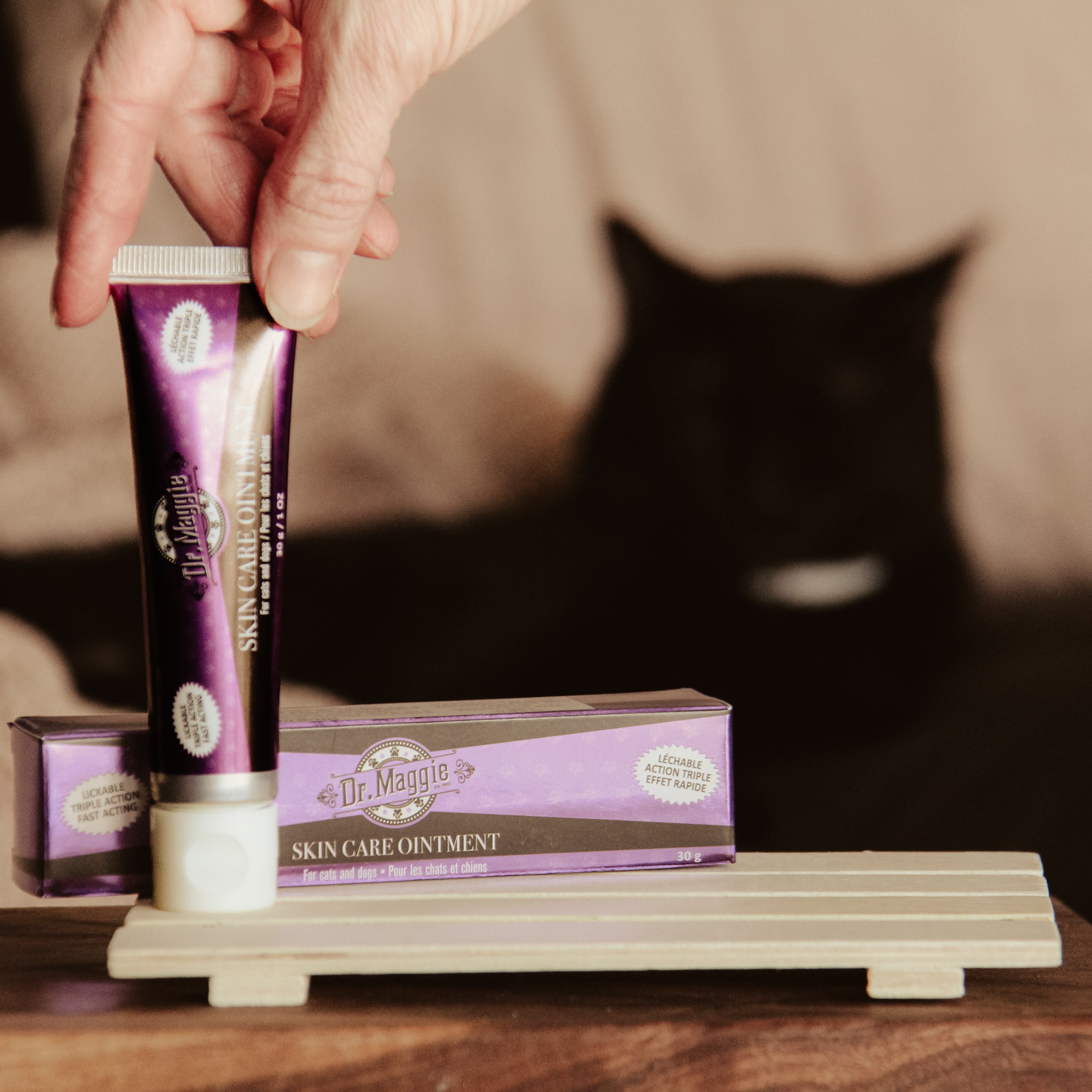 Dr. Maggie skin care ointment tube and packaging on a wooden stand with a black cat in the background.