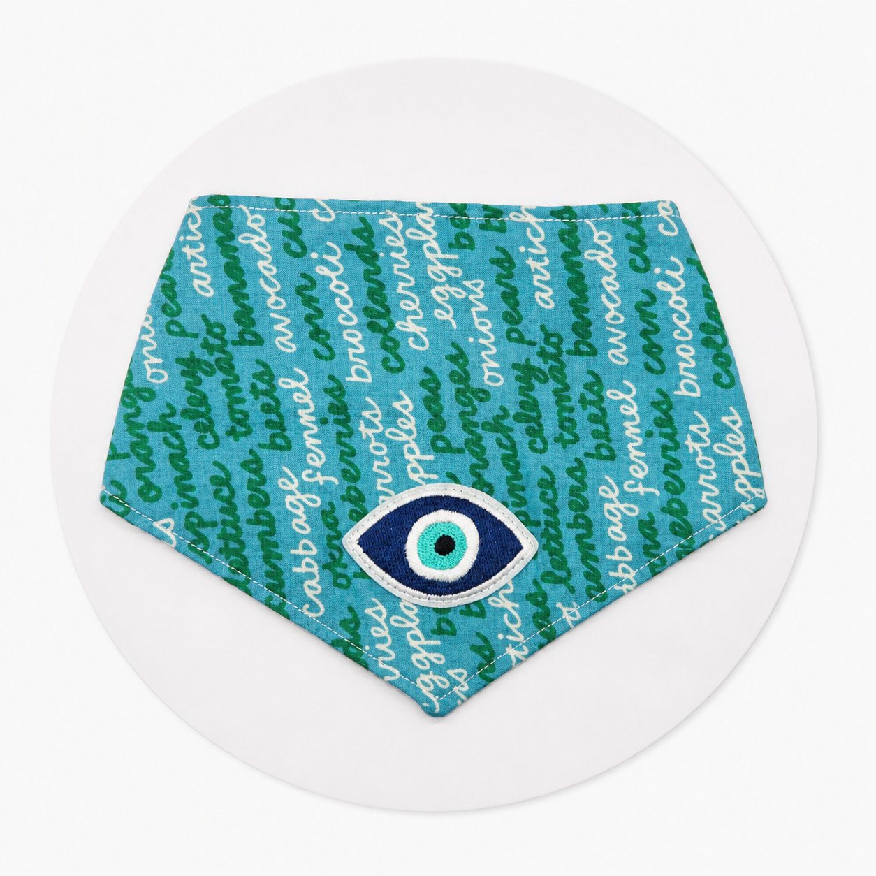 A triangular bandana-style bib or pet bandana featuring a teal and green pattern of broccoli and cabbage names, with a large embroidered evil eye motif in blue, white, and green at the center, displayed on a clean white background.