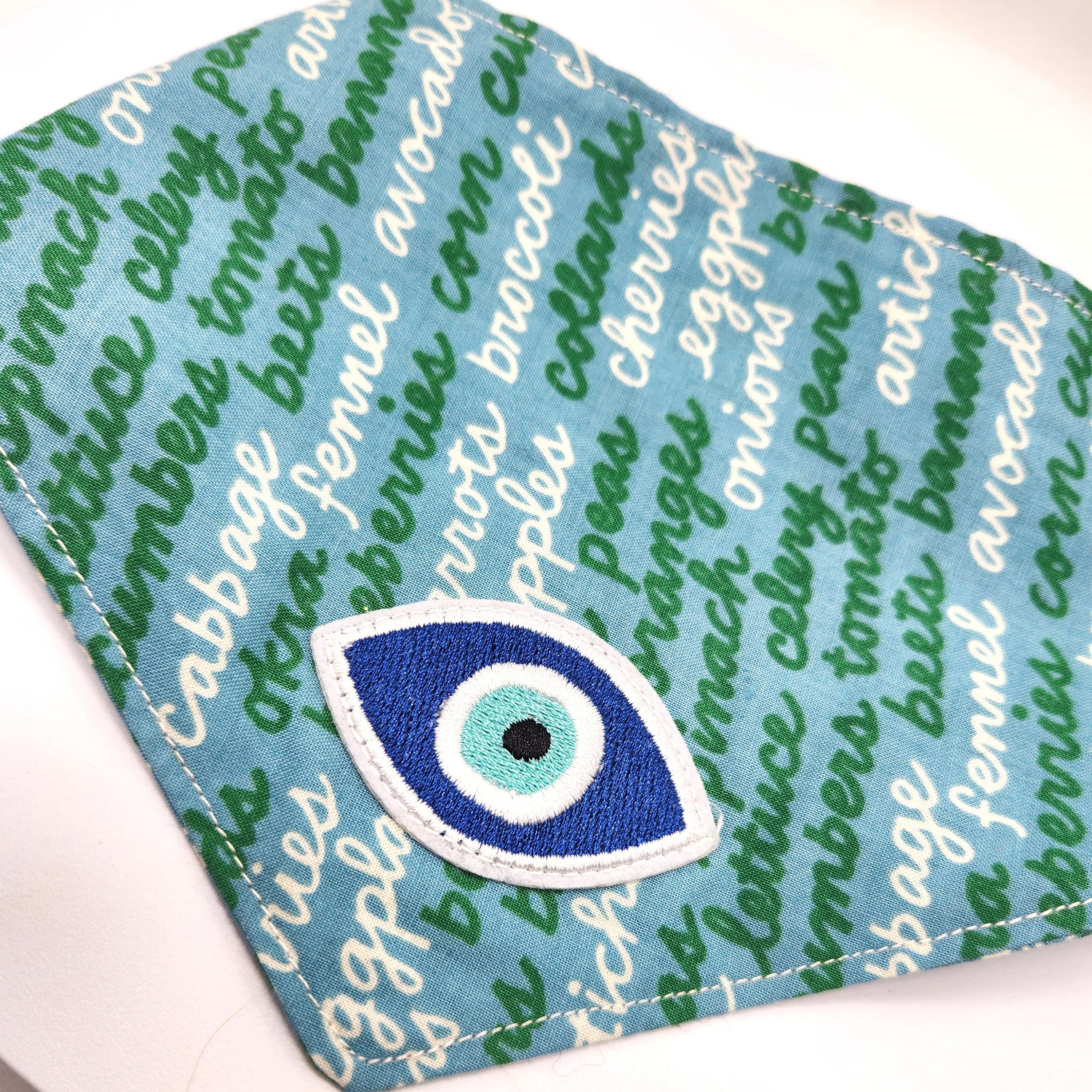 A close-up of a blue bandada for dogs, featuring a green script pattern and a prominent blue and white embroidered evil eye patch on the front, set against a white background.