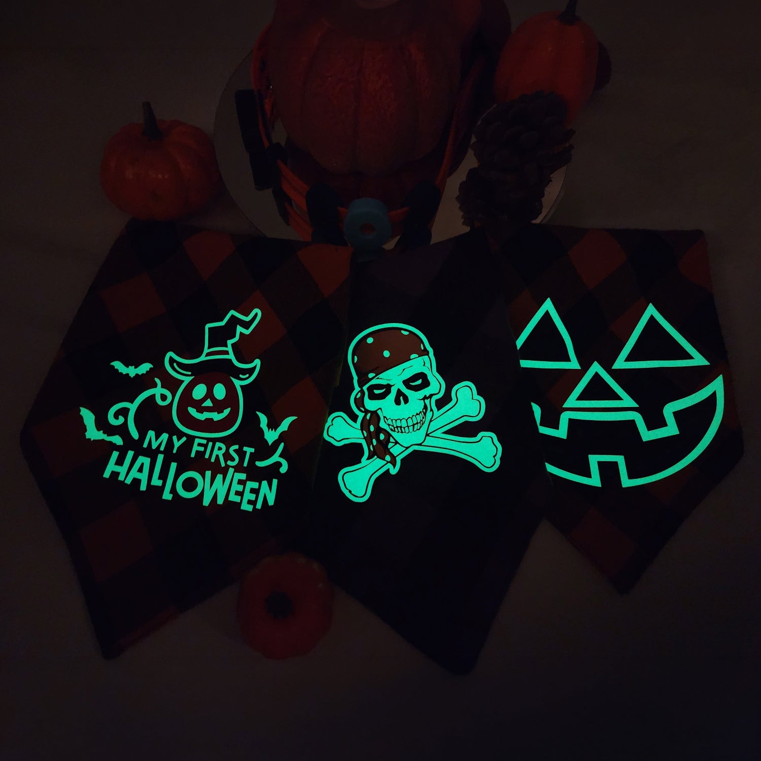 Three black plaid Halloween-themed bandanas displayed on a dark surface, glowing with green luminescent graphics: a jack-o'-lantern with the text 'MY FIRST HALLOWEEN,' a skull and crossbones with a pirate, and a jack-o'-lantern face. Pumpkins and dark decor are visible in the background.
