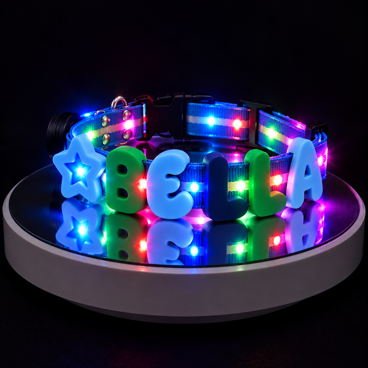 A customizable LED pet collar displayed on a circular turntable. The collar features a black plastic buckle, a black mesh band with colorful RGB LED lights, and custom light-up lettering spelling 'BELLA' with a star accent.