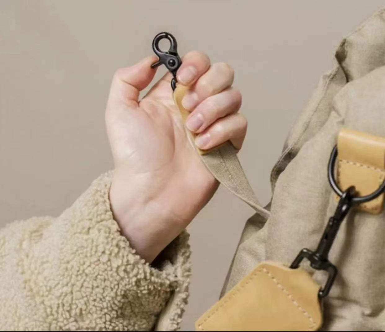 Hand holding a beige leather strap with a keychain attached, wearing a beige coat.